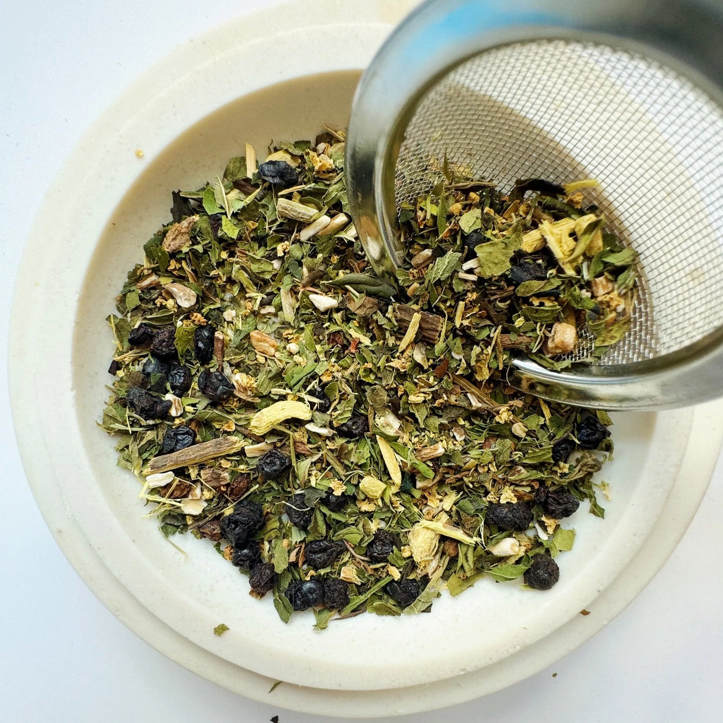 This image shows a close-up of Passage Digestive Elixir Cirene loose-leaf tea, featuring a blend of dried herbs and botanicals in a white bowl with a tea infuser. Ingredients appear to include dried berries and various green leaves, ideal for digestive support.